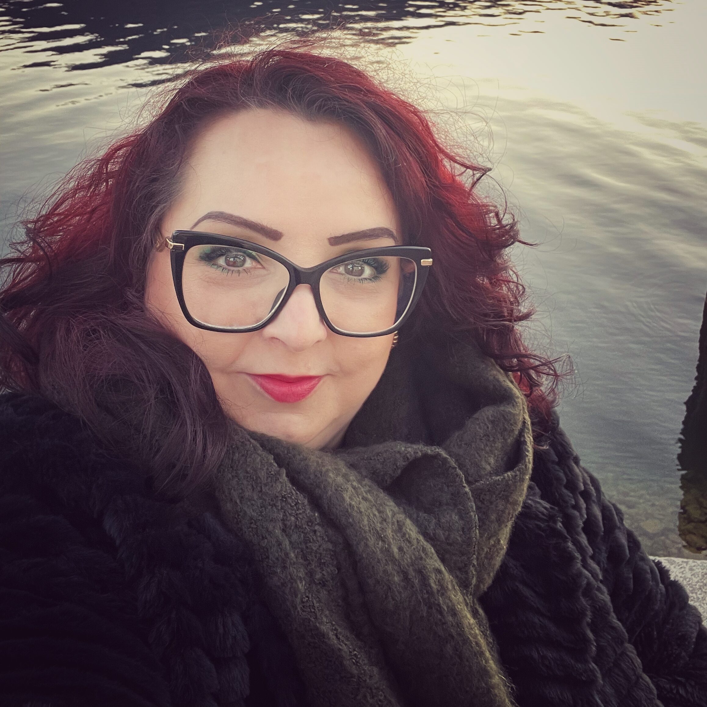Selfie-Porträt von Susan Sedlick vor einem Gewässer, im Abendlicht. Sie hat rotgefärbte Haare und eine Brille auf, und kuckt energisch-freundlich in die Kamera.